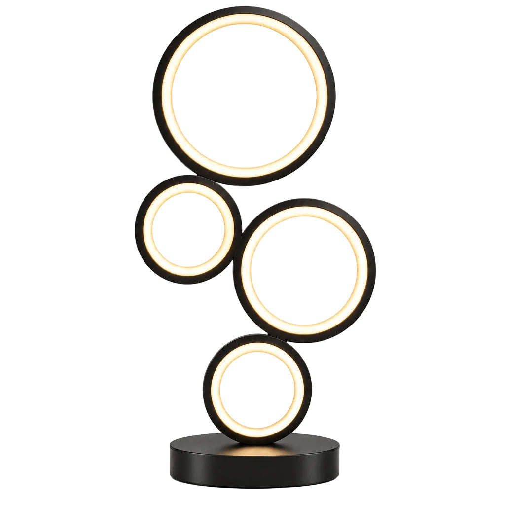 A modern table lamp with five interlocking circular light panels in black, glowing with warm white light on a wooden table. A yellow flower pot and a white mug with a yellow cat face are placed on either side of the lamp against a plain beige wall.