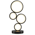 A modern table lamp with five interlocking circular light panels in black, glowing with warm white light on a wooden table. A yellow flower pot and a white mug with a yellow cat face are placed on either side of the lamp against a plain beige wall.