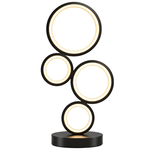 A modern table lamp with five interlocking circular light panels in black, glowing with warm white light on a wooden table. A yellow flower pot and a white mug with a yellow cat face are placed on either side of the lamp against a plain beige wall.